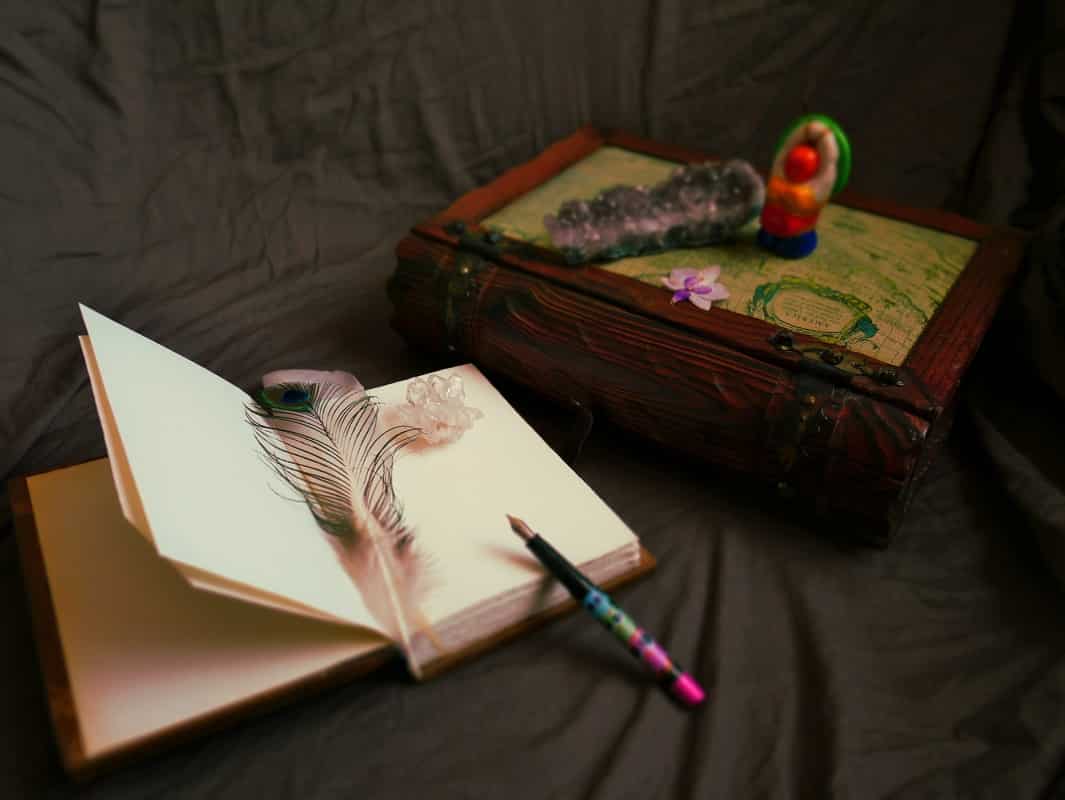 Sur un fond marron, un gros livre en bois est posé, fermé. Sur lui sont placé un amas d'améthyste, une petite fleur séchée d'orchidée, et une figurine de déesse-mère colorée. Devant, un autre livre est ouvert. Sur ses pages, blanches, sont posées deux plumes, une d'oie et une de paon, ainsi qu'un cristal de roche. Un stylo plume noir et rose est posé en équilibre sur le bord du livre.