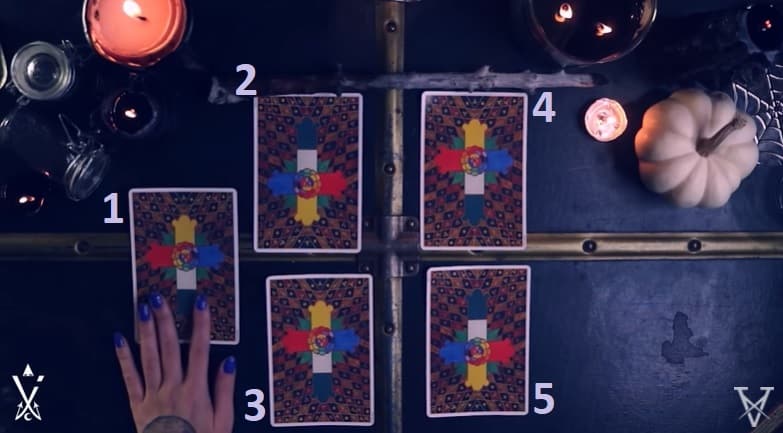 Sur une malle noire, décorée avec des bougies et une mini courge blanche, 5 cartes sont posées, en carré avec une carte seule à gauche. Une main de femme, aux ongles bleu foncé, est posée sur la carte tout à gauche.
