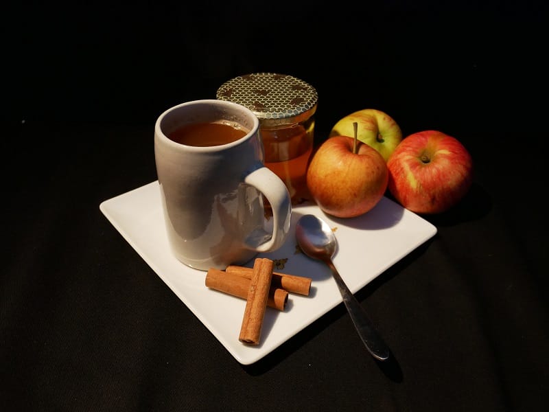 Sur fond noir, dans une assiette carrée blanche, sont posés un pot de miel, une tasse risse remplie d’un liquide brun, trois bâtons de cannelle, une cuillère en inox et une pomme. Deux autres pommes sont posées contre celle-ci, hors de l’assiette.