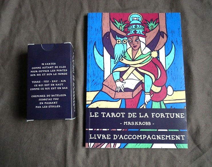 Sur un tissu gris sont posés à gauche le boîtier cartonné bleu-violet sombre (on voit sa face arrière avec du texte) et à droite le livret accompagnement du Tarot de la Fortune.