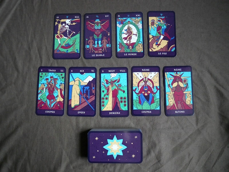 Sur un tissu gris sont posées deux séries de cartes, face visible. En haut, de gauche à droite on a : l’arcane 13 sans nom, Le Diable, Le Monde et Le Fou. En bas, de gauche à droite on a : le 3 de Coupes, le 6 d’Épées, le 9 de Deniers, la Reine de Coupes et la Reine de Bâtons. Tout en bas est posé le reste des cartes en paquet, dos visible.