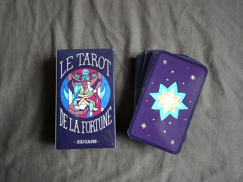 Sur un tissu gris, on voit à gauche un boîtier de Tarot, bleu-violet sombre avec au centre un détail d’une carte et le titre du jeu : « Le Tarot de la Fortune ». A droite est posé un tas de carte, dont on voit le dos, violet avec une grosse étoile jaune et bleu, ainsi que des petites étoiles jaunes.