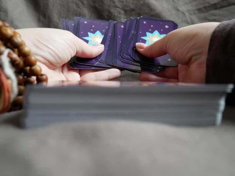 Sur un tissu gris, deux mains tiennent quelques cartes, vues de dos. Au premier plan, un peu flou, on voit un paquet de cartes.