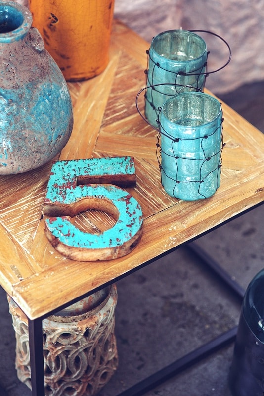 Sur une table en bois sont posés des éléments de décoration : de gauche à droite, un vase en terre cuite, un gros 5 en bois, et deux photophores entouré d’un maillage grossier, le tout peint en turquoise.