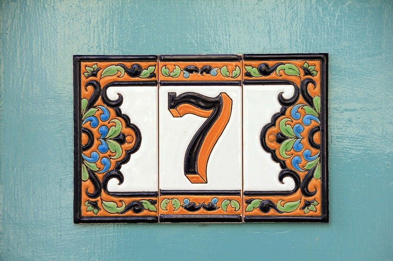 Plaque tripartite en faïence portant le numéro 7, orange et noir avec touches de bleu et de vert, sur fond blanc, et fixée sur un mur bleu.