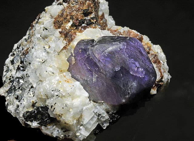 Saphir brut, violet, sur cristaux de feldspath (albite) et de mica (biotite) donnant une mâcle blanche avec traces noires et rouille.