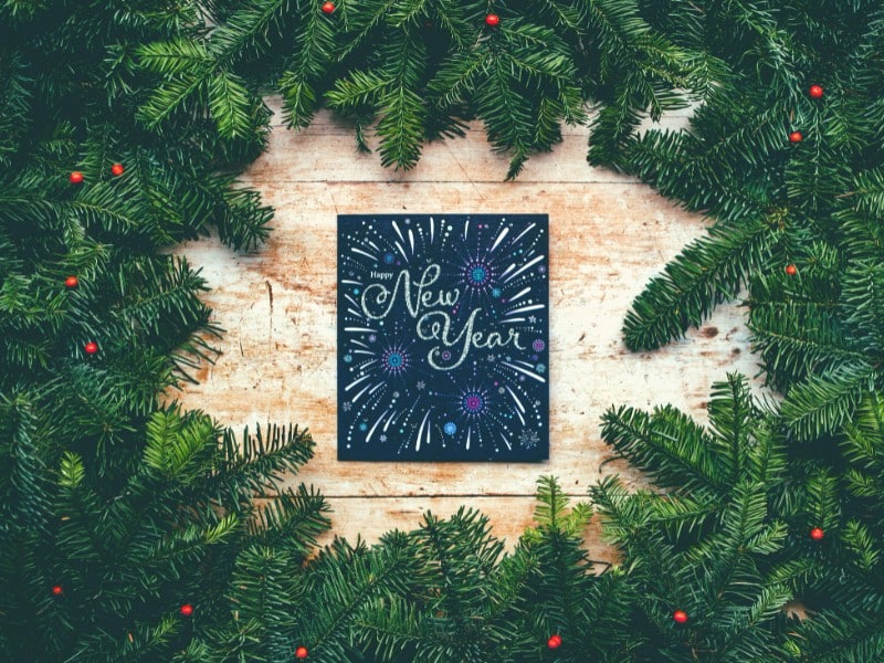 Une carte en papier noir, avec écrit « Happy New Year » et comportant des dessins de feux d’artifice, est posée sur une table en bois clair et entourée de branches de sapins avec quelques baies rouges.