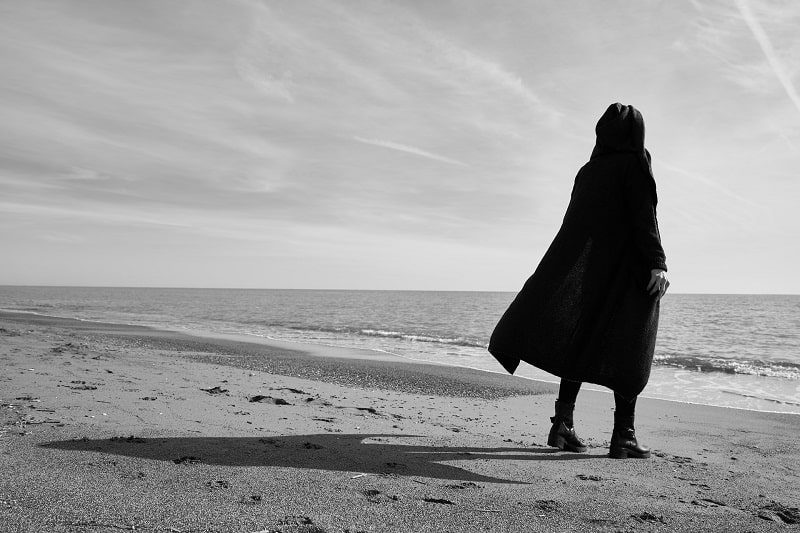 En noir et blanc, sur une plage, une personne habillée en noir avec un long manteau et la capuche sur la tête se tient debout à droite, de dos.