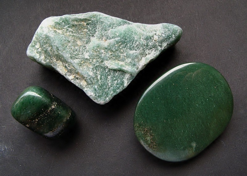 Trois aventurines sur fond noir, avec de gauche à droite : une pierre roulée vert sombre, un morceau brut vert clair, et un cabochon vert sombre.