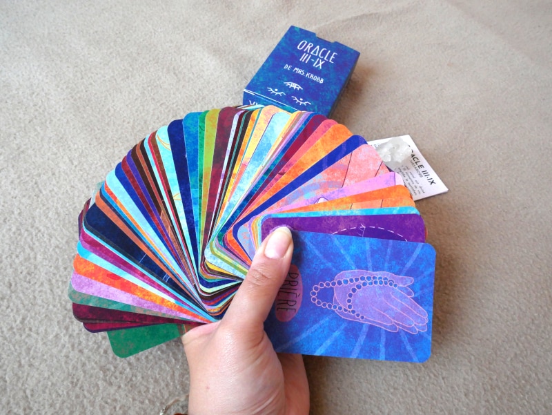 Main présentant le deck en entier, en éventail, faces visibles, pour bien montrer les nuances de couleurs. Derrière on voit le livret et le boîtier du jeu.