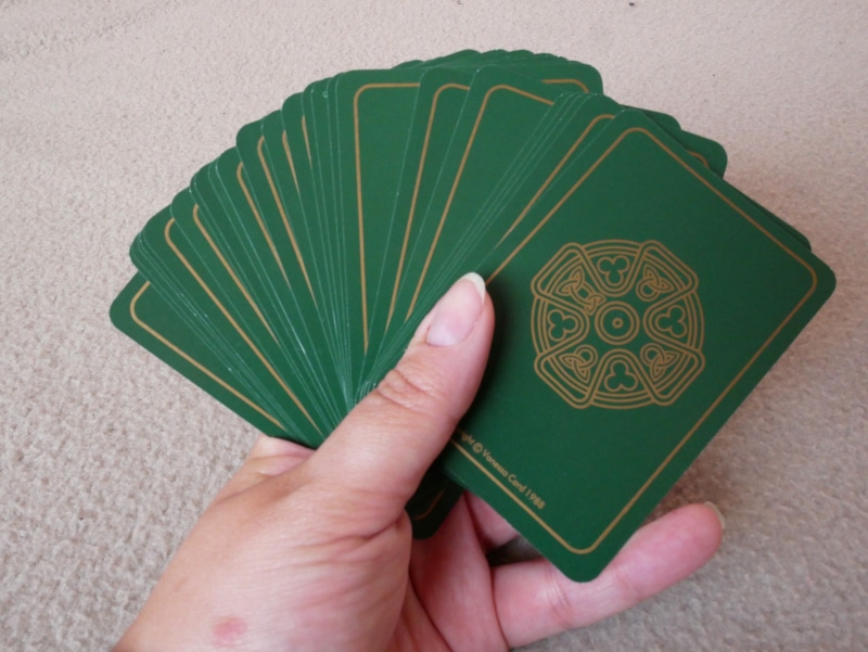 Main tenant le paquet de carte en éventail, dos des cartes visibles. Le dos des cartes est vert, avec un liseré doré et une croix celte dorée au centre.