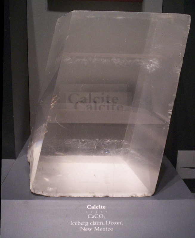 Grand cristal optique sur son présentoir de musée. On peut voir l’effet optique sur le nom « calcite » derrière le cristal.