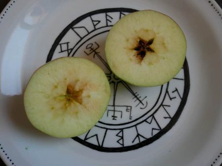 Deux moitiés de pommes, coupée horizontalement, et montrant le pentagramme formé par les pépins. La pomme est posée sur une assiette blanche peinte en noir avec un Vegvisir entouré du Futhark.